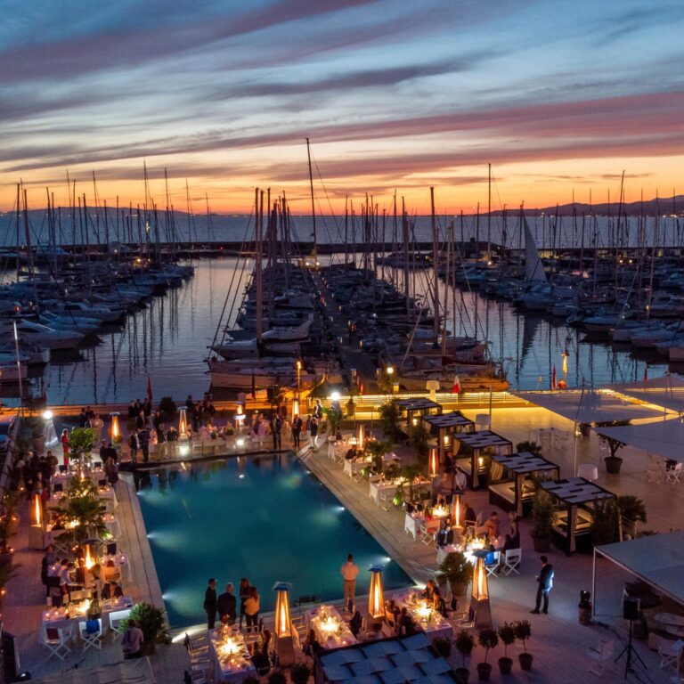Vista aerea notturna del Marina Club Scarlino con la piscina illuminata e tavoli eleganti allestiti per un evento aziendale, circondati dal porto e dagli yacht al tramonto.