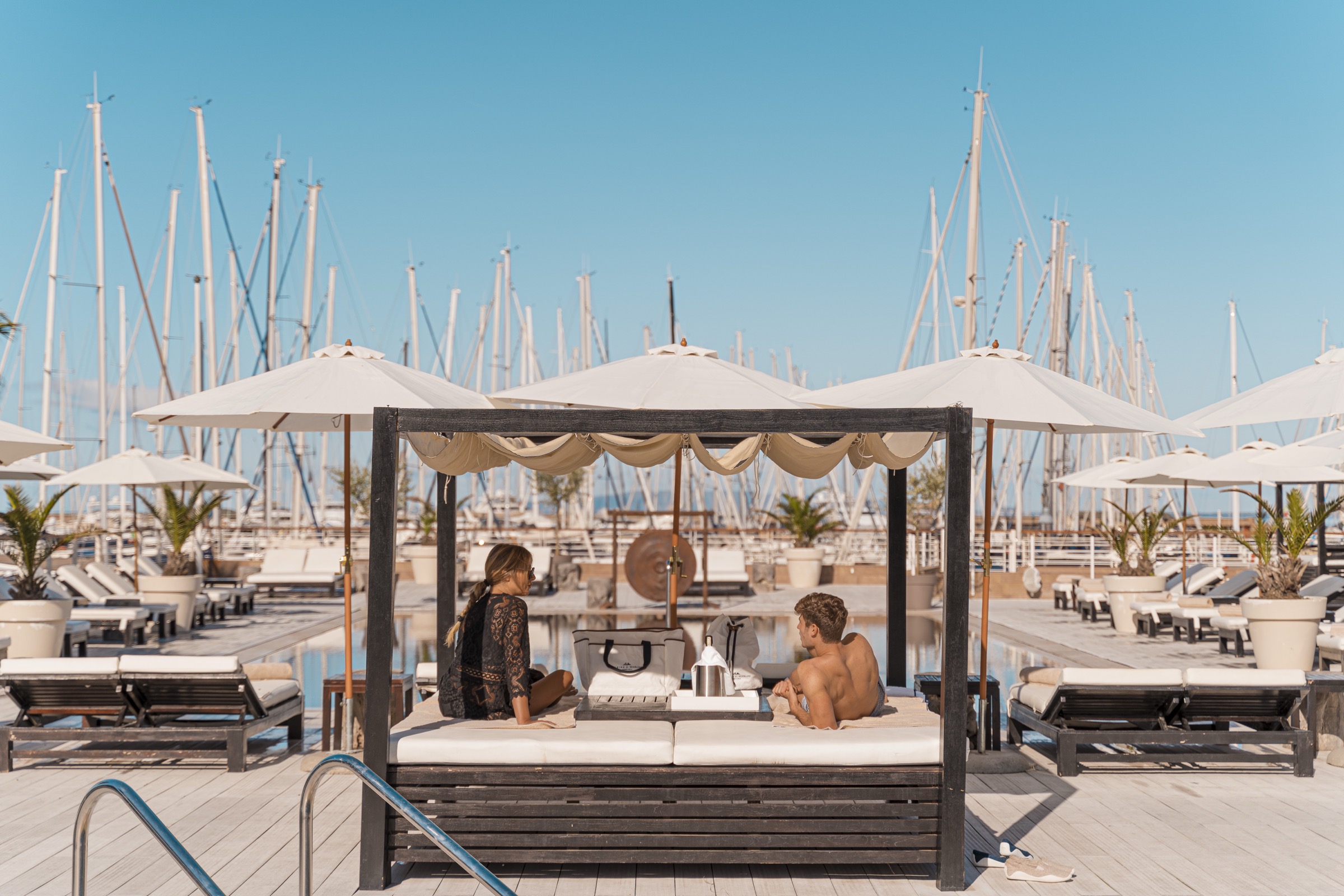 Coppia in relax su daybed a bordo piscina presso lo Yacht Club & Pool del Marina di Scarlino Resort con sfondo di alberi di barche a vela.