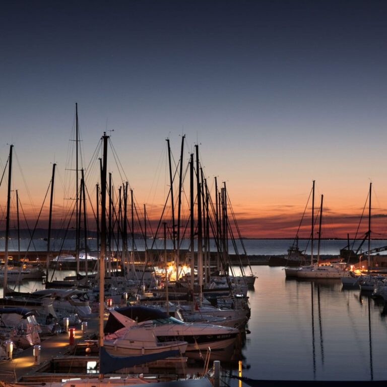 Barche a vela e yacht ormeggiati con cura nei moli del Marina di Scarlino sotto un cielo crepuscolare arancione e blu.